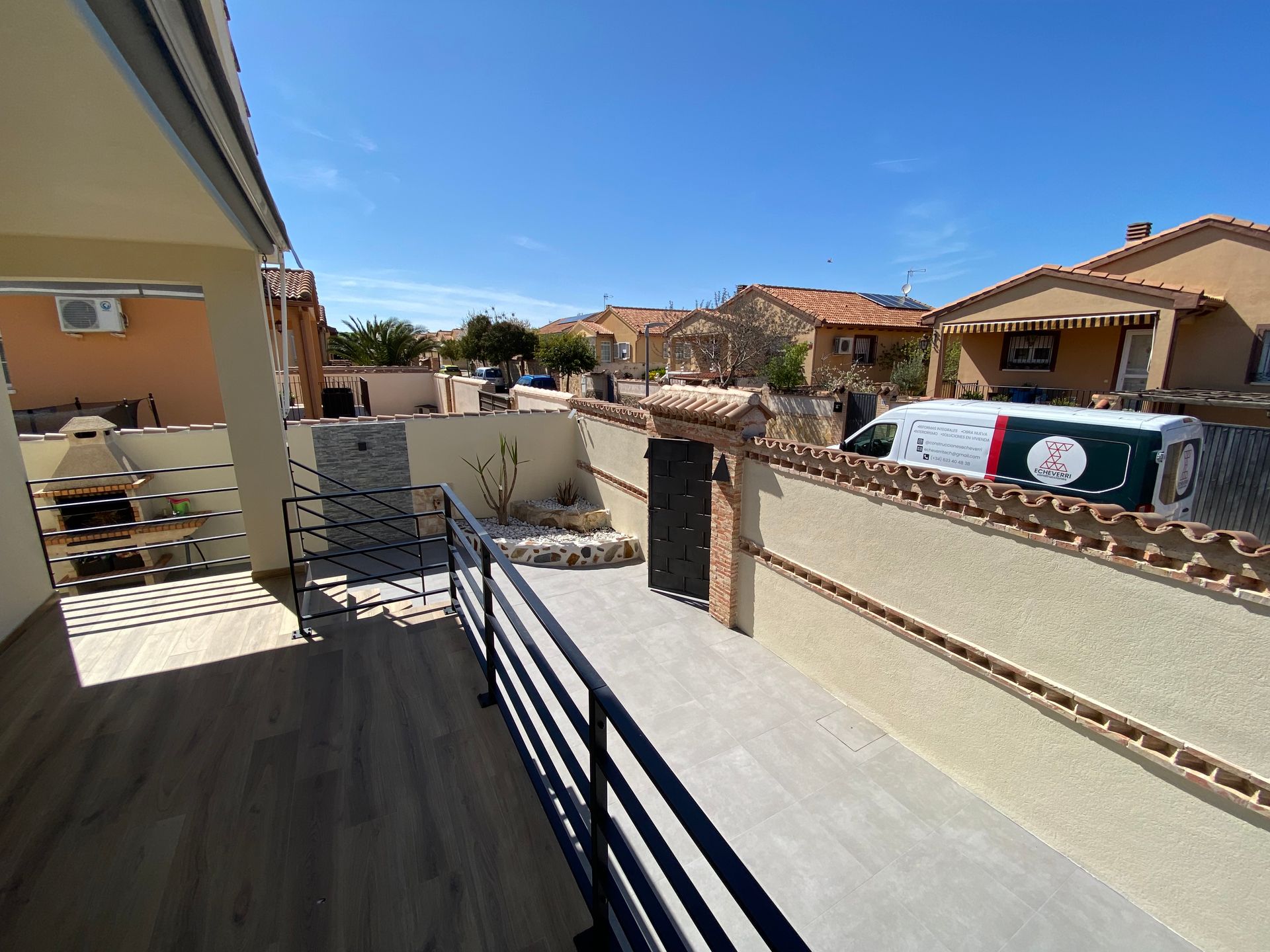Vista desde el balcón: barandilla, pasarela de hormigón, pared beige, portón, cielo azul, casas.