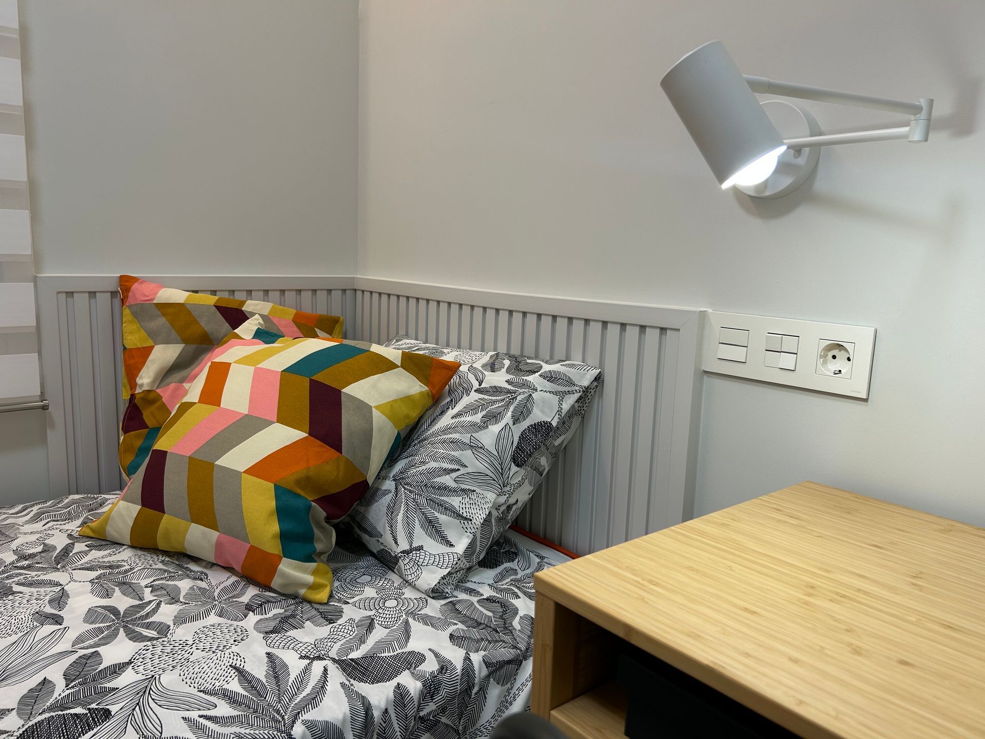 Cama con almohadas decorativas, mesita de noche y lámpara de pared.