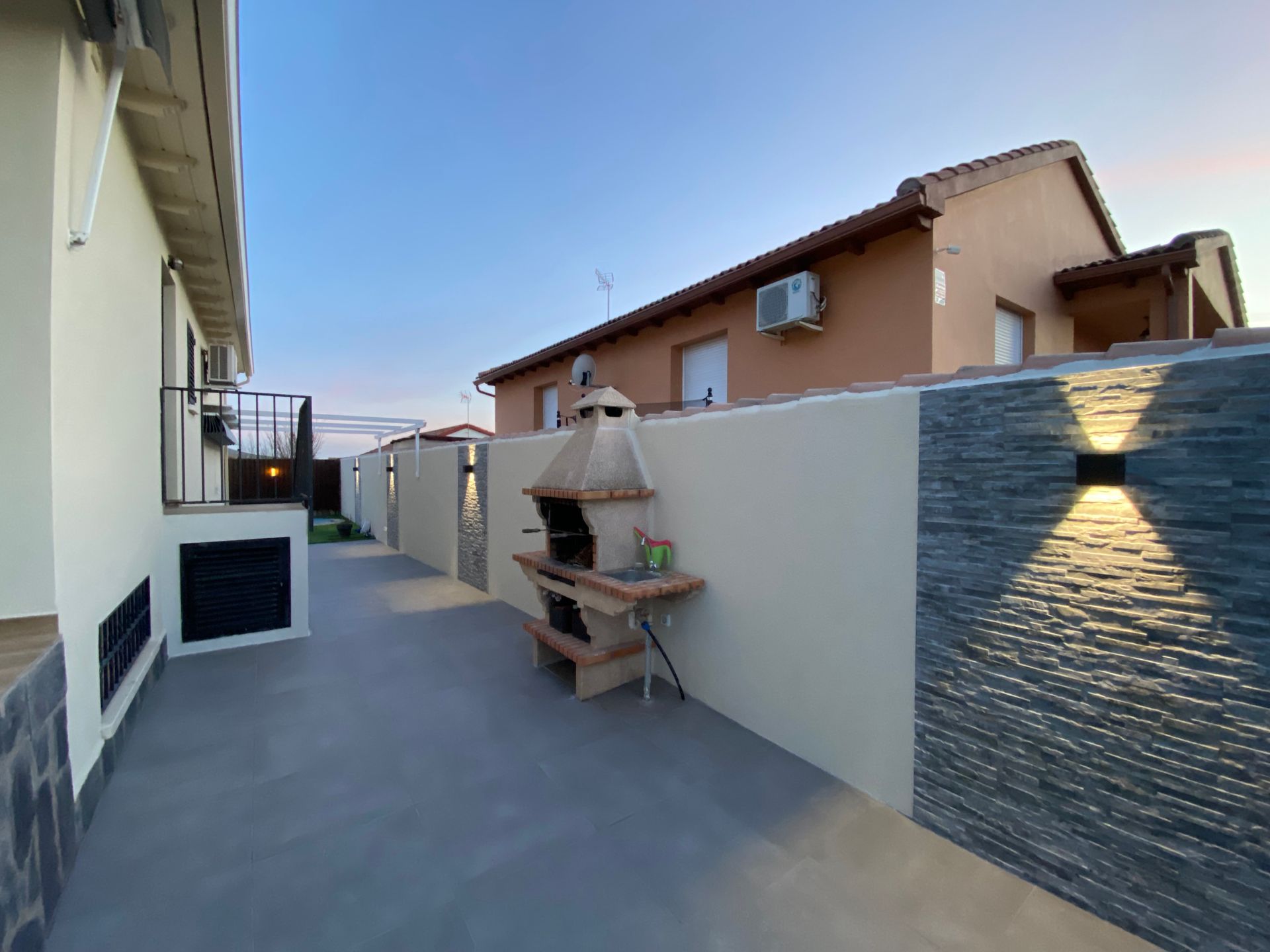 Patio trasero con parrilla y apliques de pared exterior, entre casa beige y muro alto.