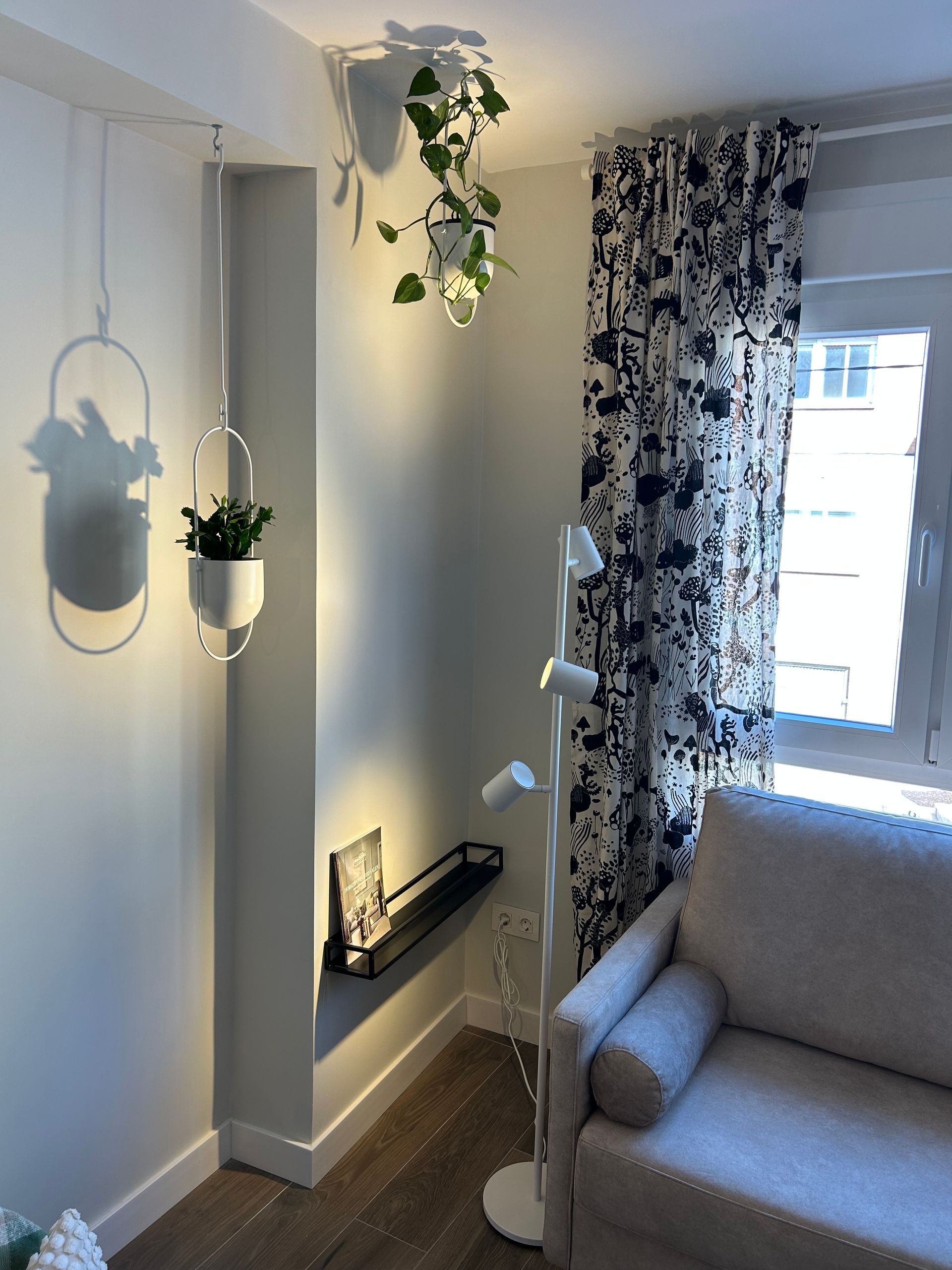 Habitación en esquina con plantas colgando del techo, una lámpara de pie y un sofá cerca de una ventana con cortinas florales.