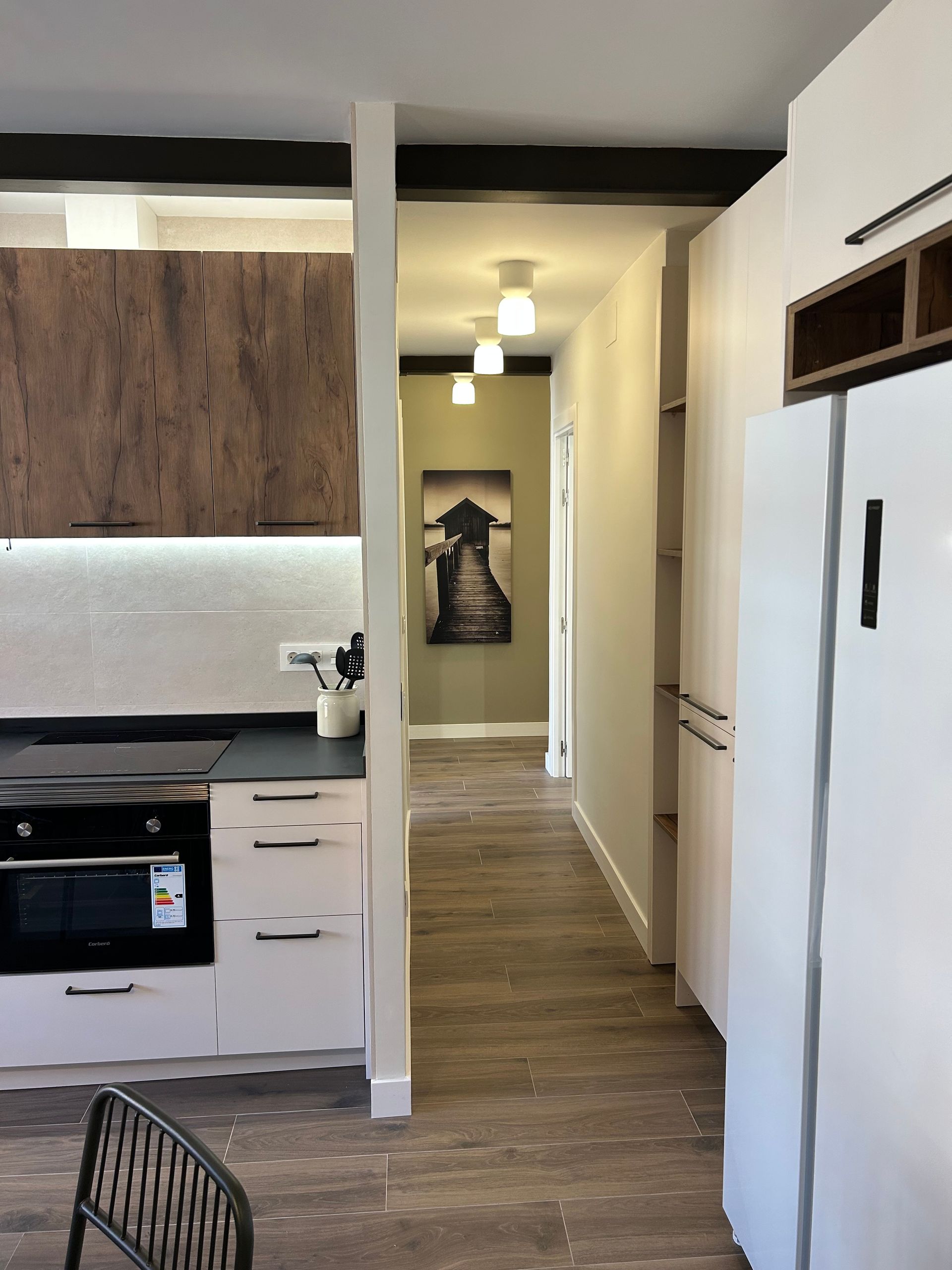 Cocina y pasillo con gabinetes blancos y de madera, encimeras oscuras y obras de arte al final del pasillo.