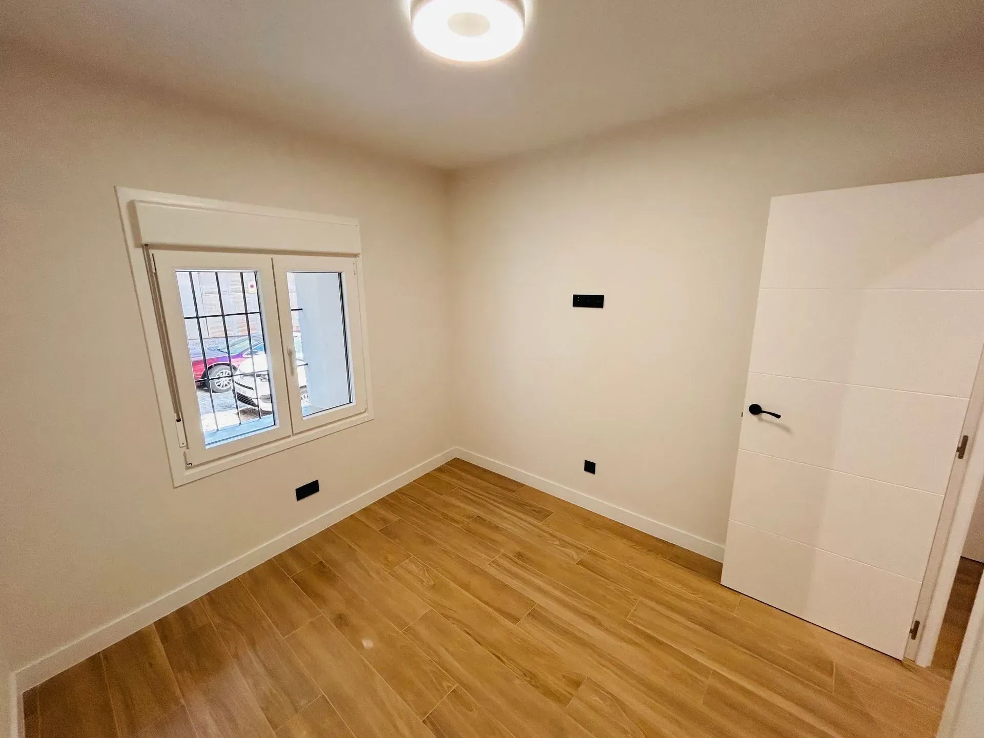 Habitación vacía con suelo de madera, paredes blancas, ventana, puerta y luz de techo.