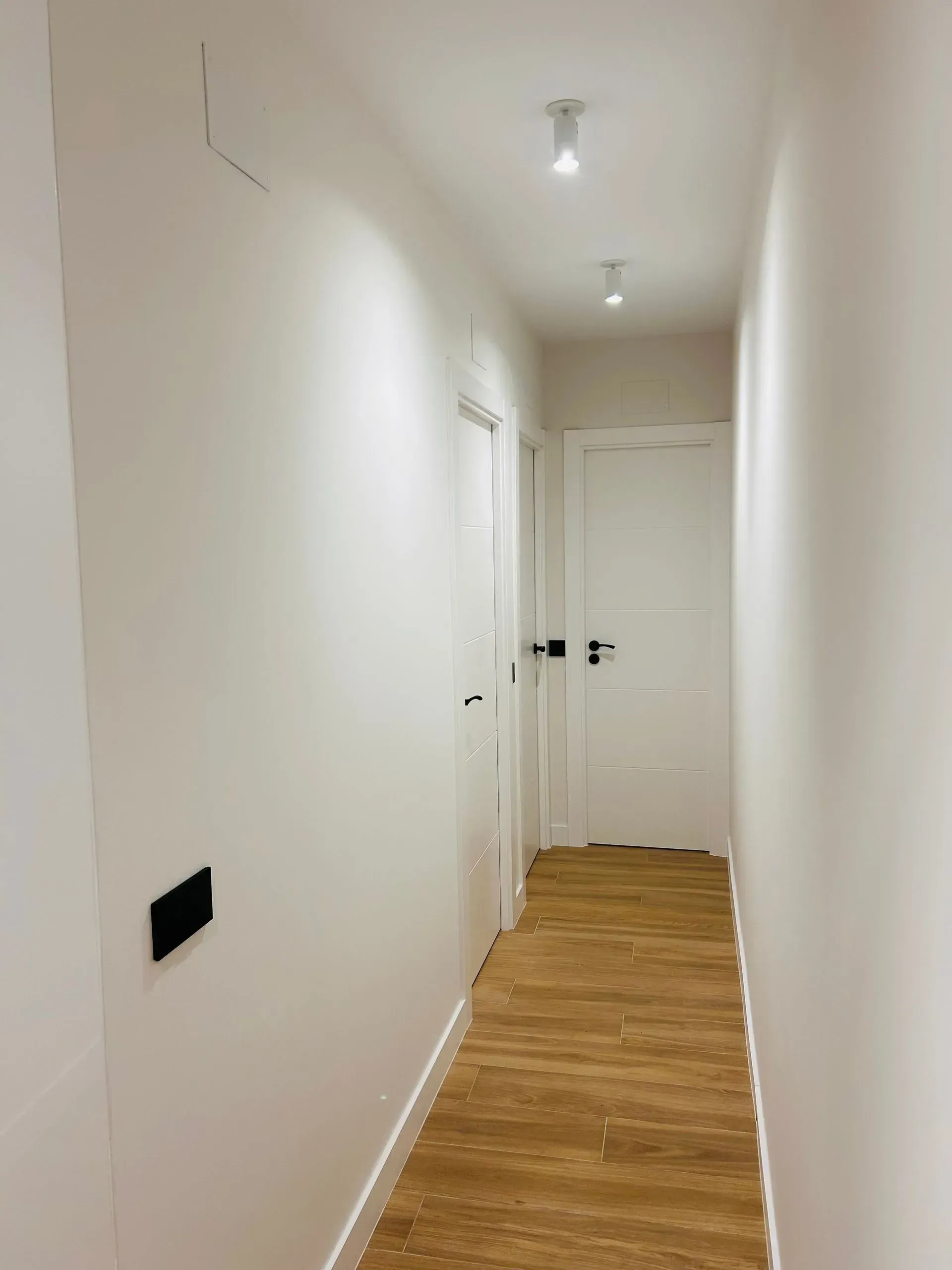 Pasillo largo con paredes blancas, tres puertas, focos y suelo de madera.