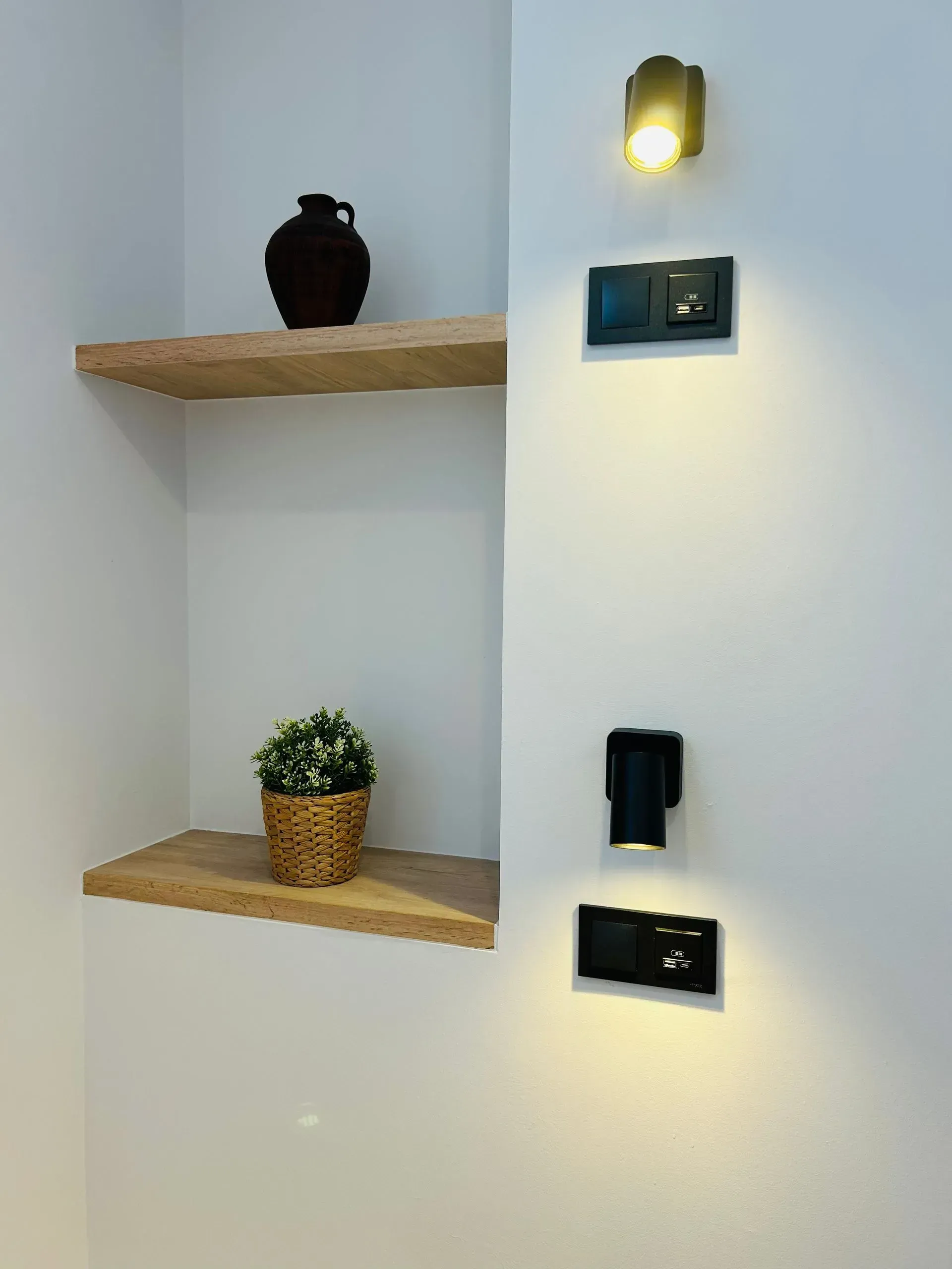 Pared blanca con estantes de madera, objetos decorativos e interruptores de luz negros. Una pequeña lámpara ilumina los interruptores.