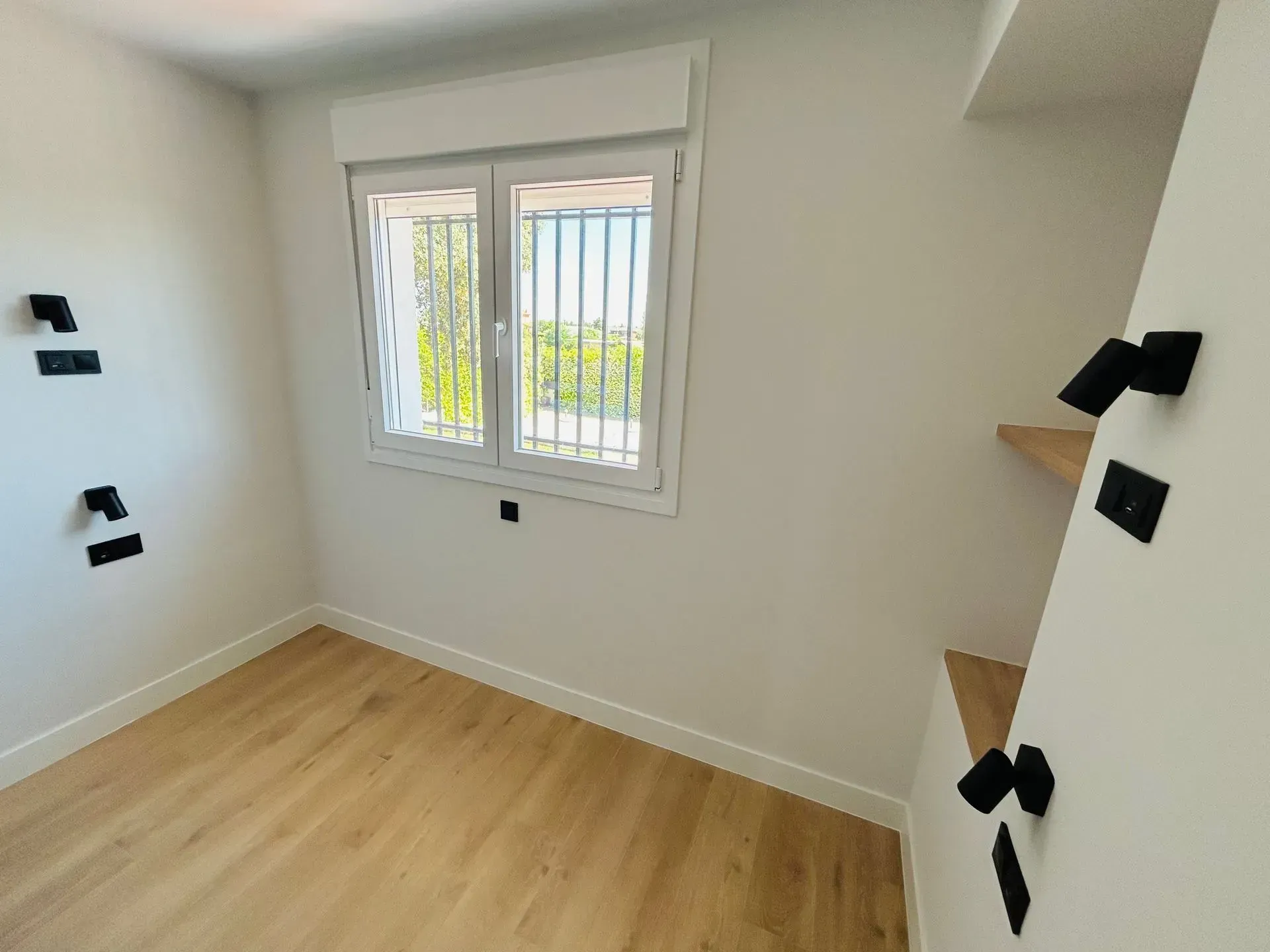 Habitación vacía con ventana, suelo de madera y paredes blancas con artefactos eléctricos negros.