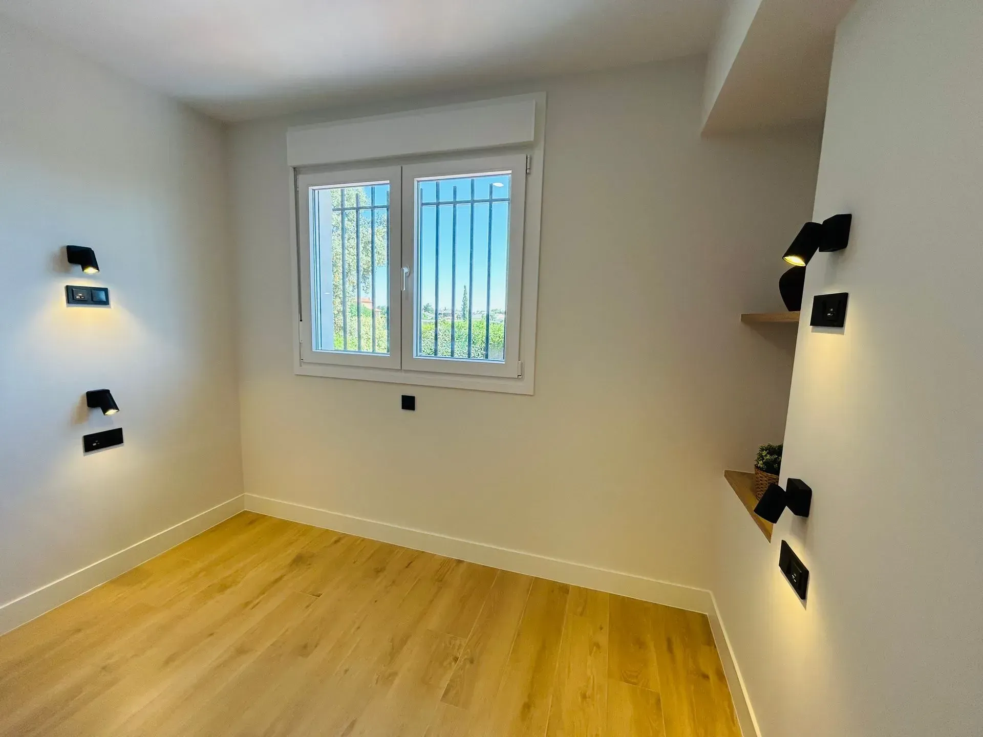 Habitación vacía con una ventana, paredes blancas, piso de madera y luces negras montadas en la pared.