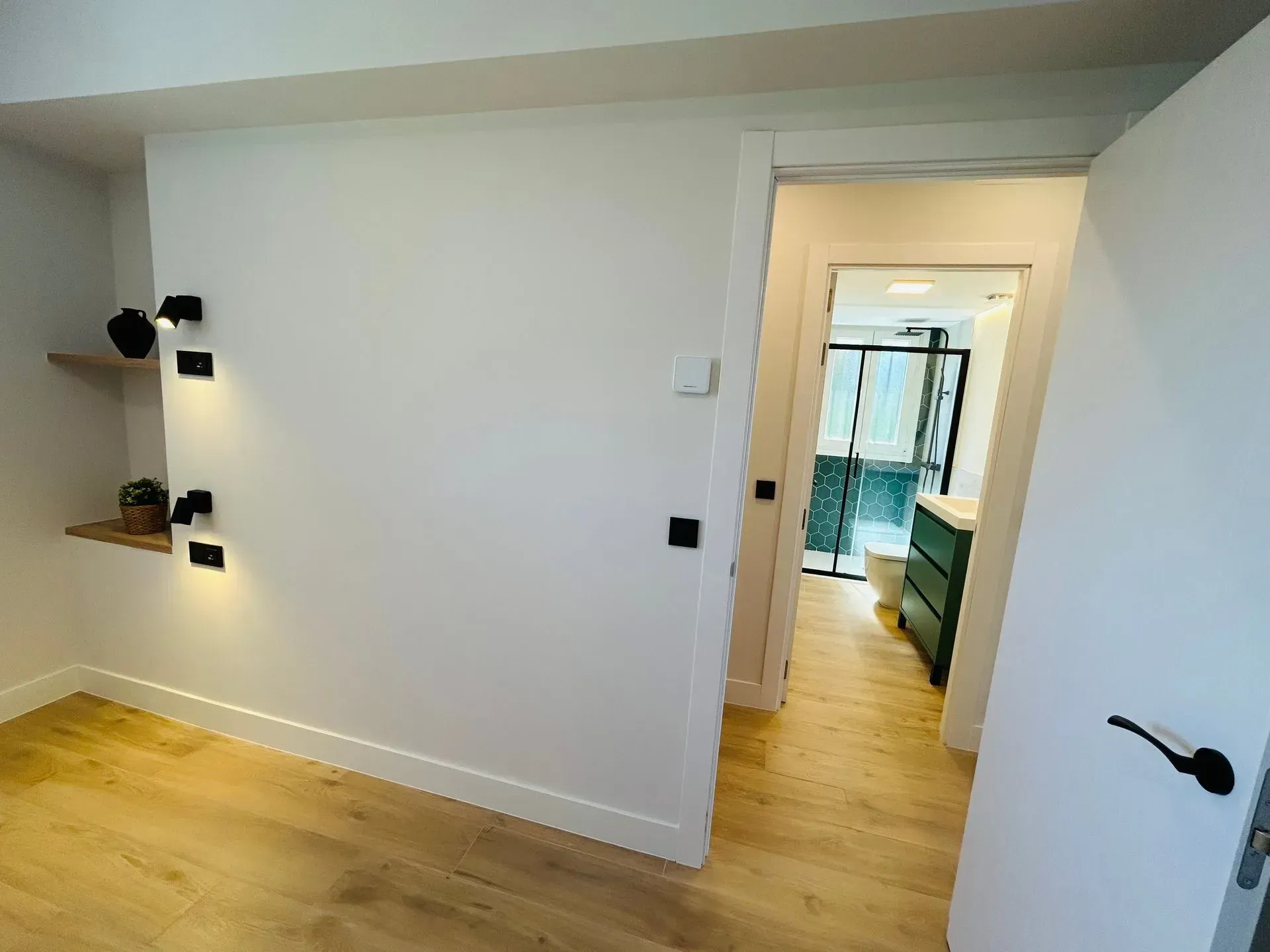 Interior de dormitorio con paredes blancas, suelo de madera y una puerta que conduce a un baño.