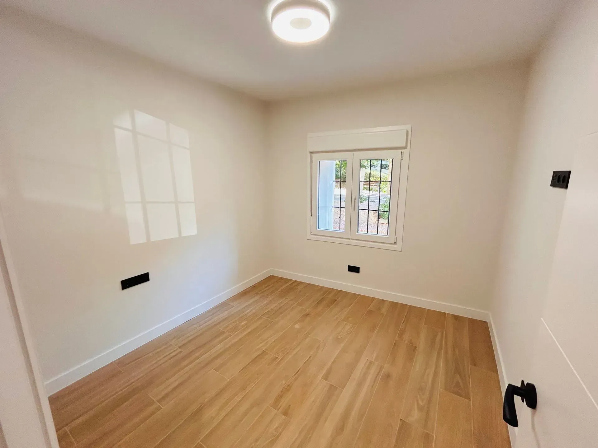Habitación vacía con suelo imitación madera, ventana con persiana y luz de techo. Paredes blancas.