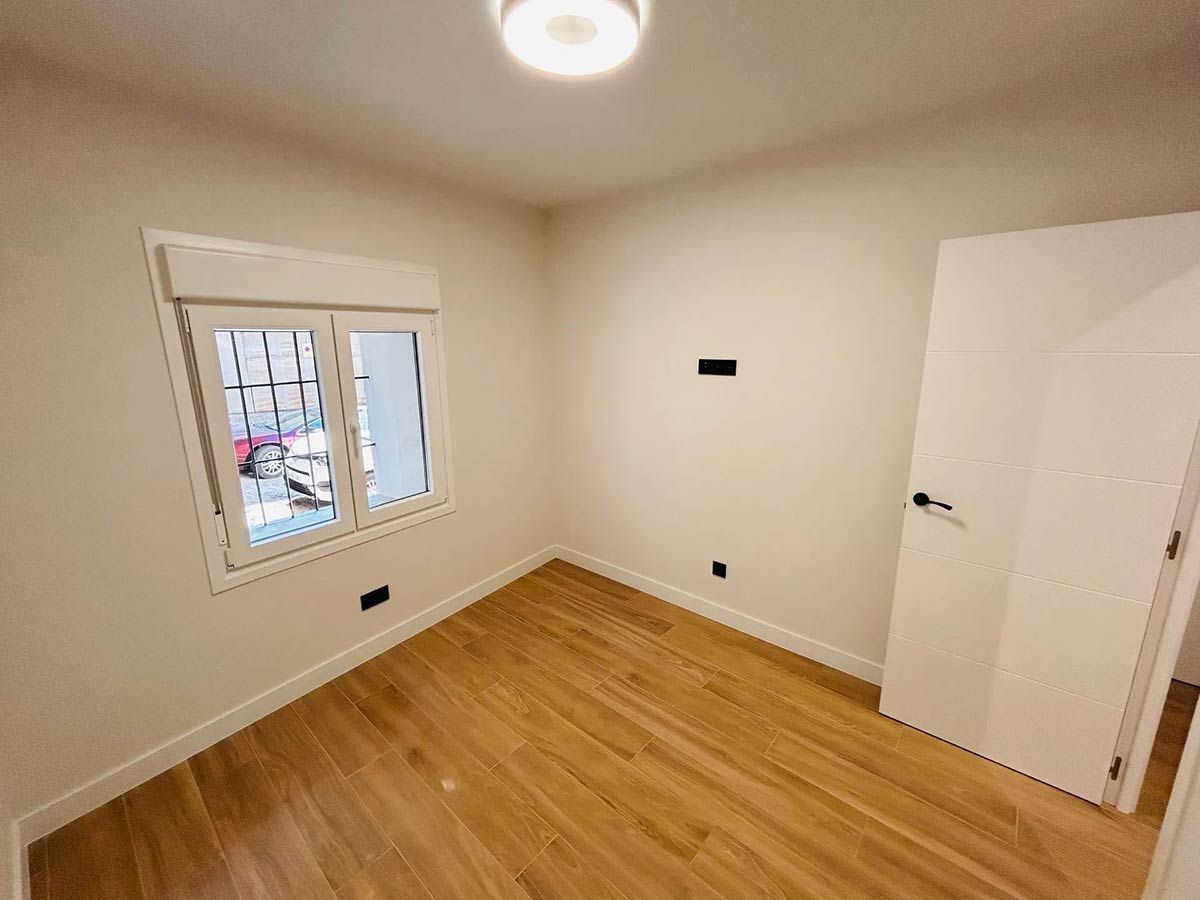 Habitación vacía con piso de baldosas con apariencia de madera, paredes blancas, ventana, puerta blanca cerrada e iluminación empotrada.