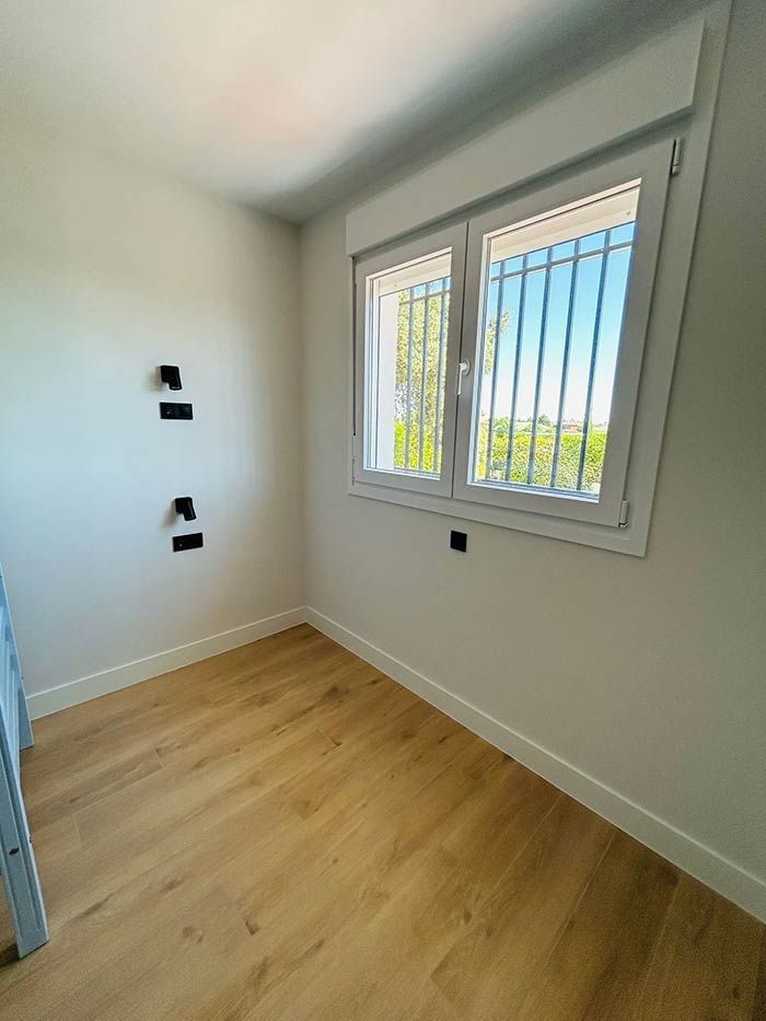 Habitación blanca vacía con pisos de madera clara, ventana con rejas y enchufes eléctricos.