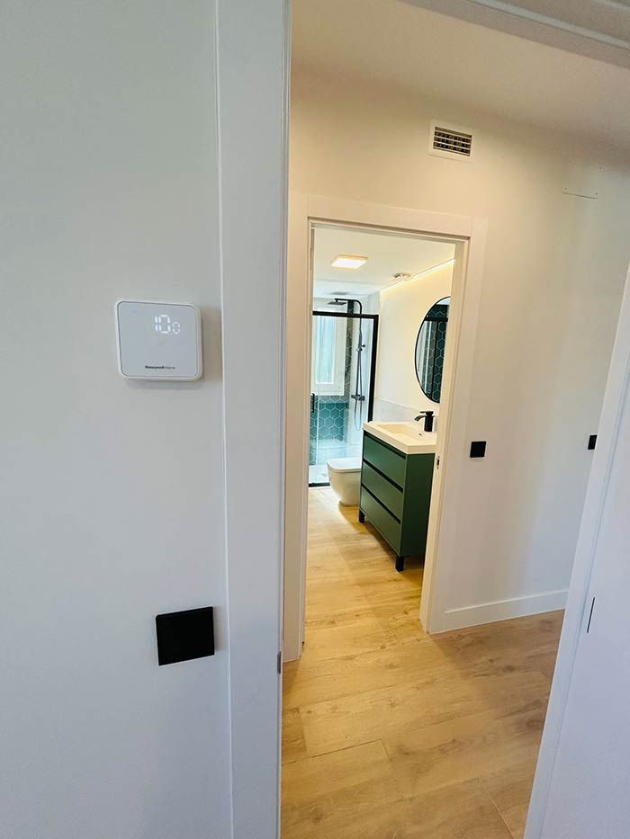 Pasillo con termostato en la pared izquierda, que conduce a un baño con tocador verde y espejo redondo.