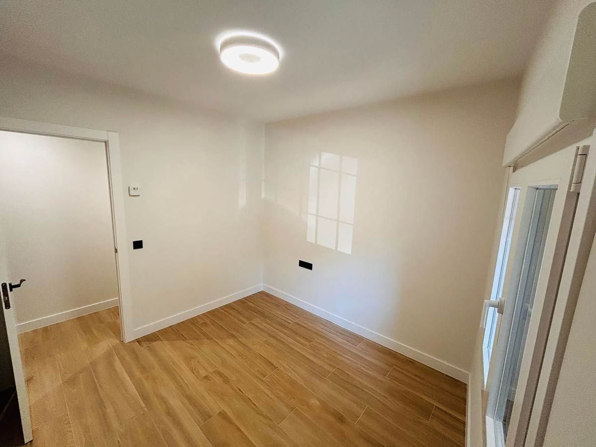 Habitación vacía con suelo de madera y paredes blancas, una puerta y una ventana.