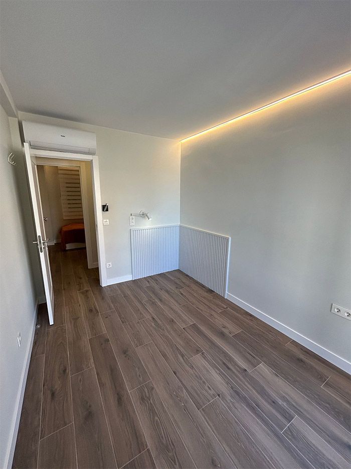 Habitación vacía con suelo de madera y paredes blancas. Una puerta da a otra habitación. Tiras de luces LED en el techo.