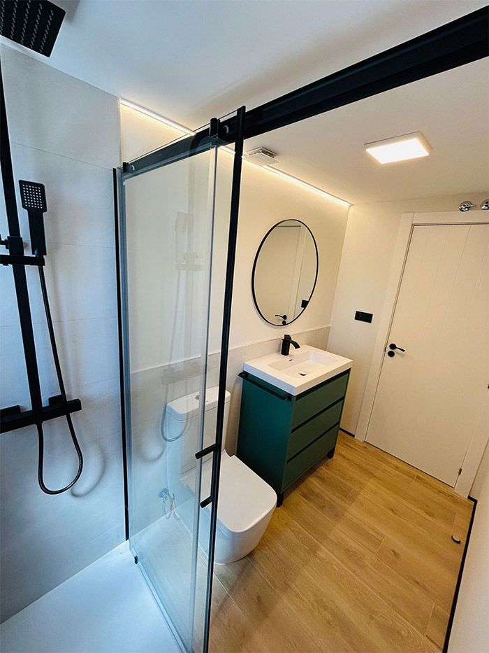 Baño moderno con ducha, inodoro y tocador. Tocador verde, grifería negra y suelo imitación madera.