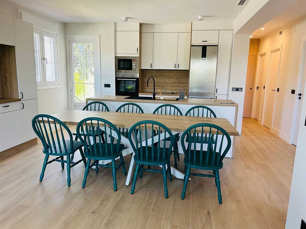 Cocina con mesa grande de madera, sillas color verde azulado, gabinetes blancos y pisos de madera.