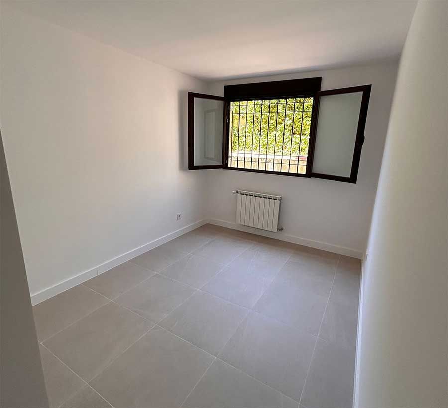 Habitación vacía con ventana, radiador y suelo de baldosas; paredes blancas.