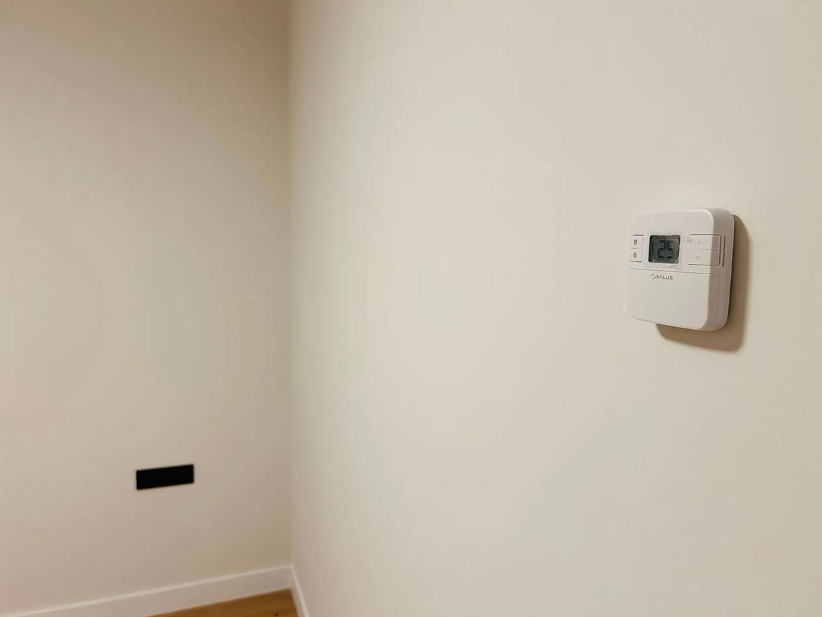 Paredes blancas con un termostato y una salida rectangular oscura cerca de la esquina de una habitación.