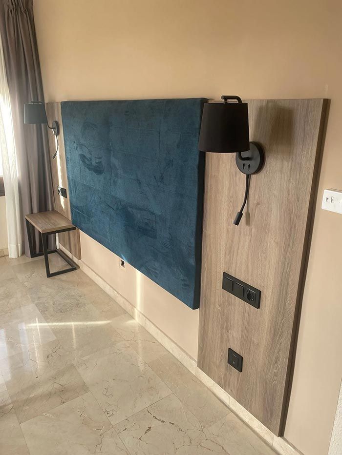 Pared de dormitorio moderna con cabecera tapizada en verde azulado, panel lateral de madera, aplique, enchufes y un pequeño banco.