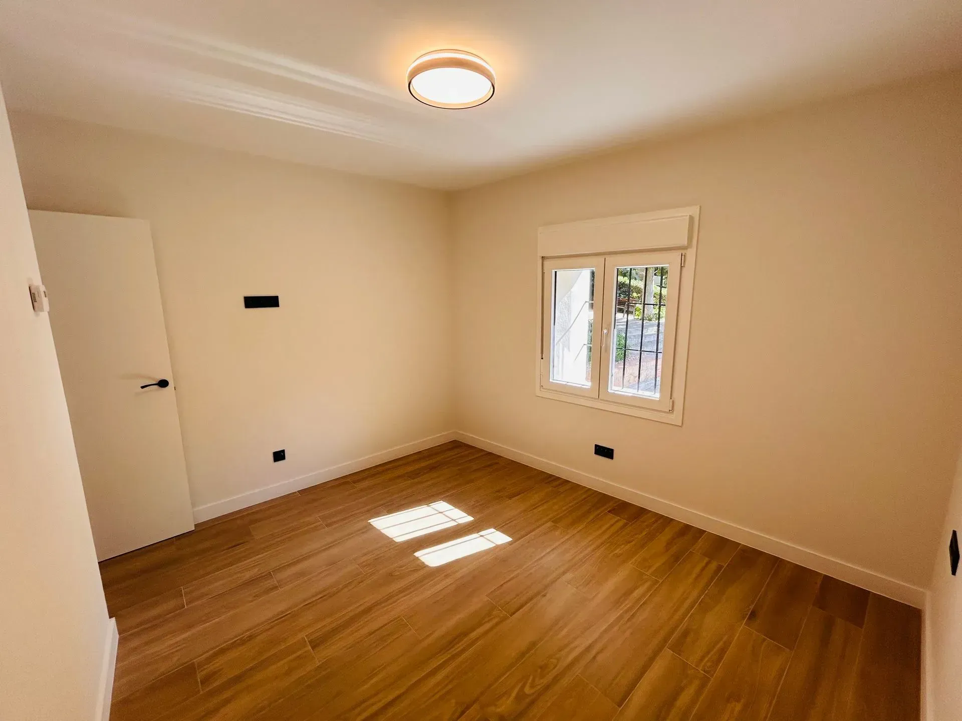 Habitación vacía con piso de madera clara, paredes blancas, puerta, ventana con persianas cerradas y luz de techo.