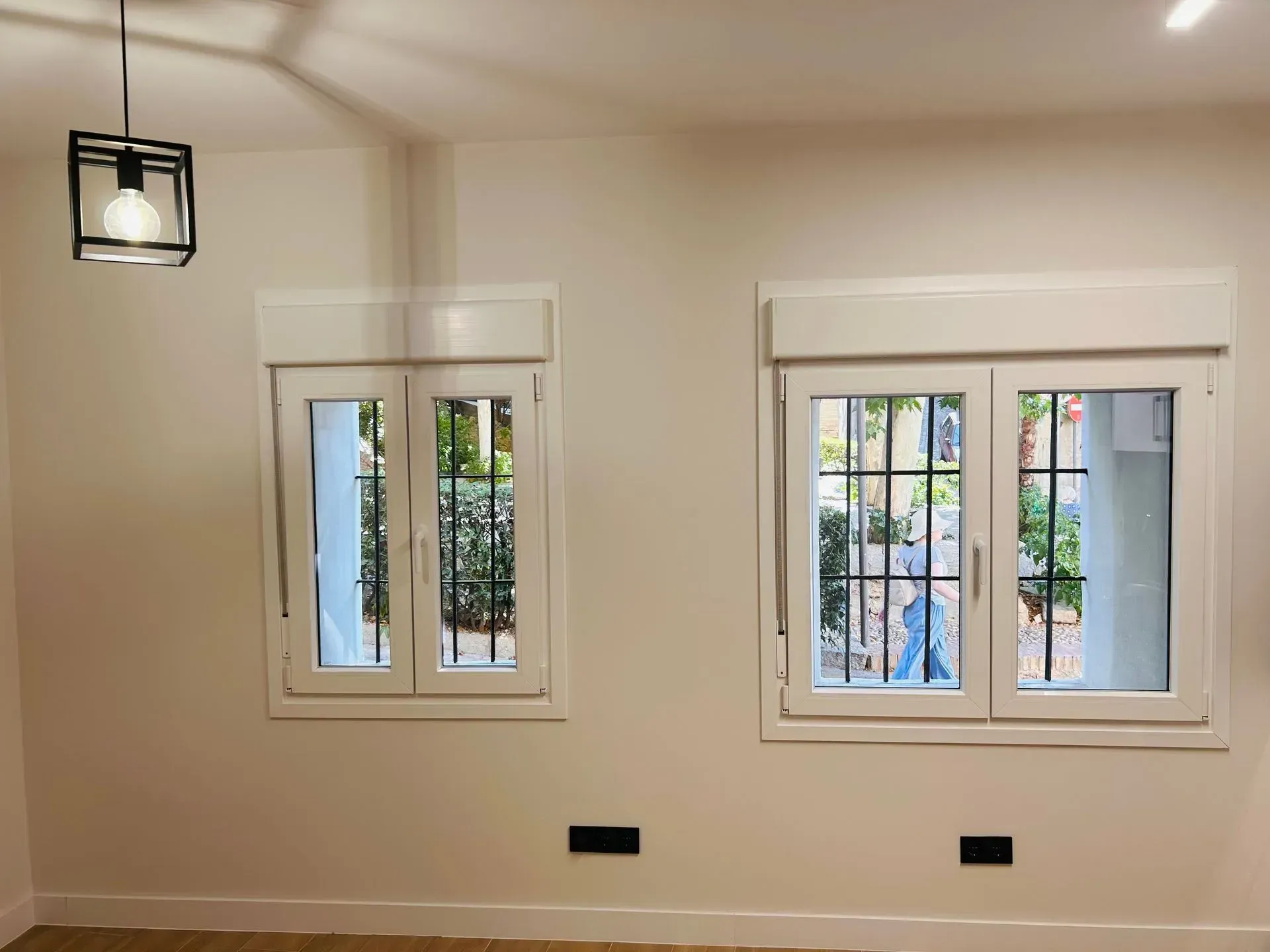 Dos ventanas con marco blanco y rejas de seguridad negras. También se ven paredes beige, una lámpara moderna y dos enchufes negros.
