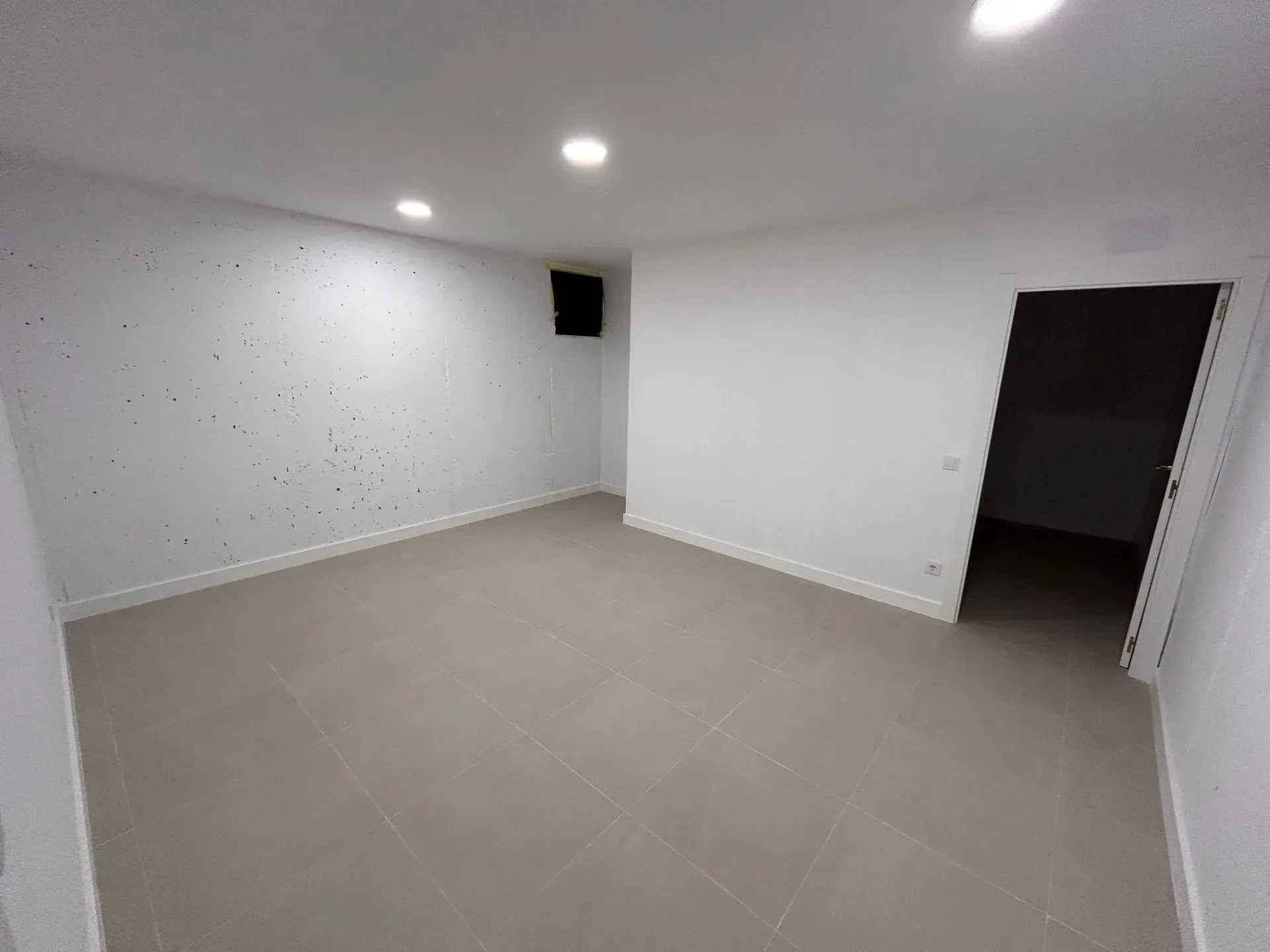 Habitación vacía, de paredes blancas, con suelo de baldosas grises y una puerta. Iluminación empotrada en el techo.