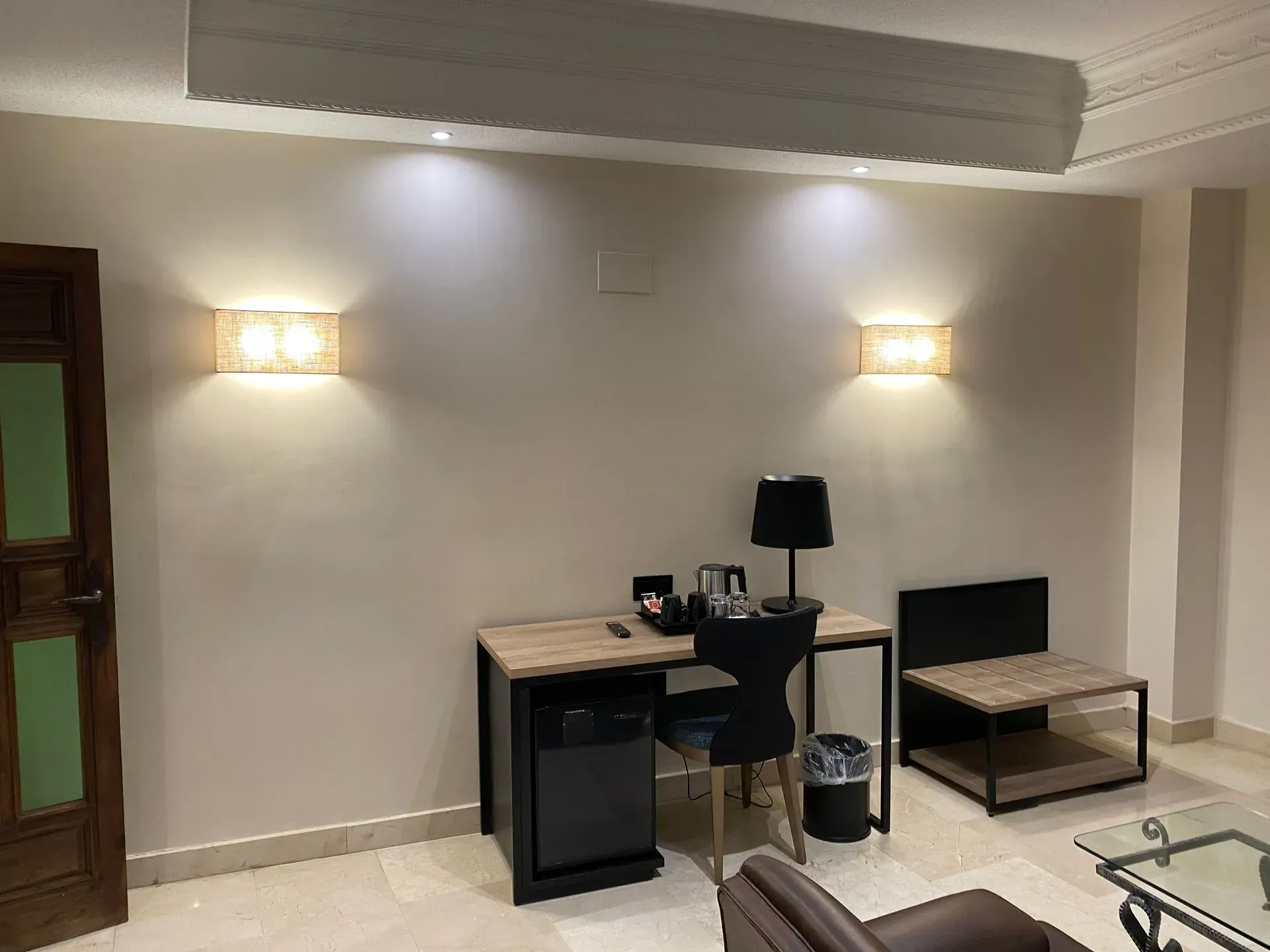 Interior de habitación de hotel con escritorio, silla y mesita auxiliar. Dos apliques de pared y una puerta oscura. Paredes y suelo beige.