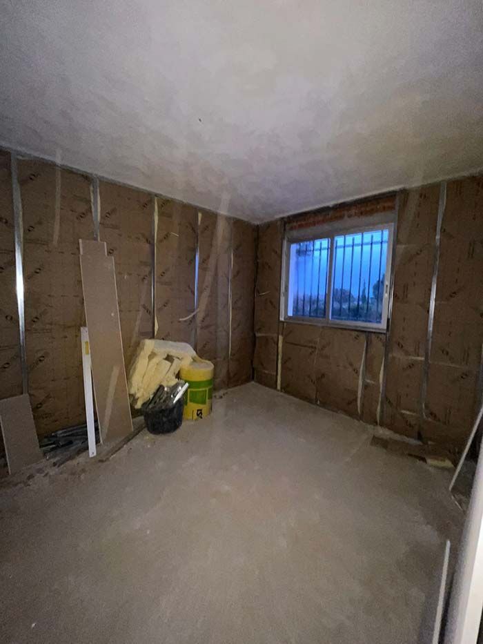 Interior de una habitación en construcción con aislamiento expuesto, ventana con rejas de seguridad y piso de concreto.