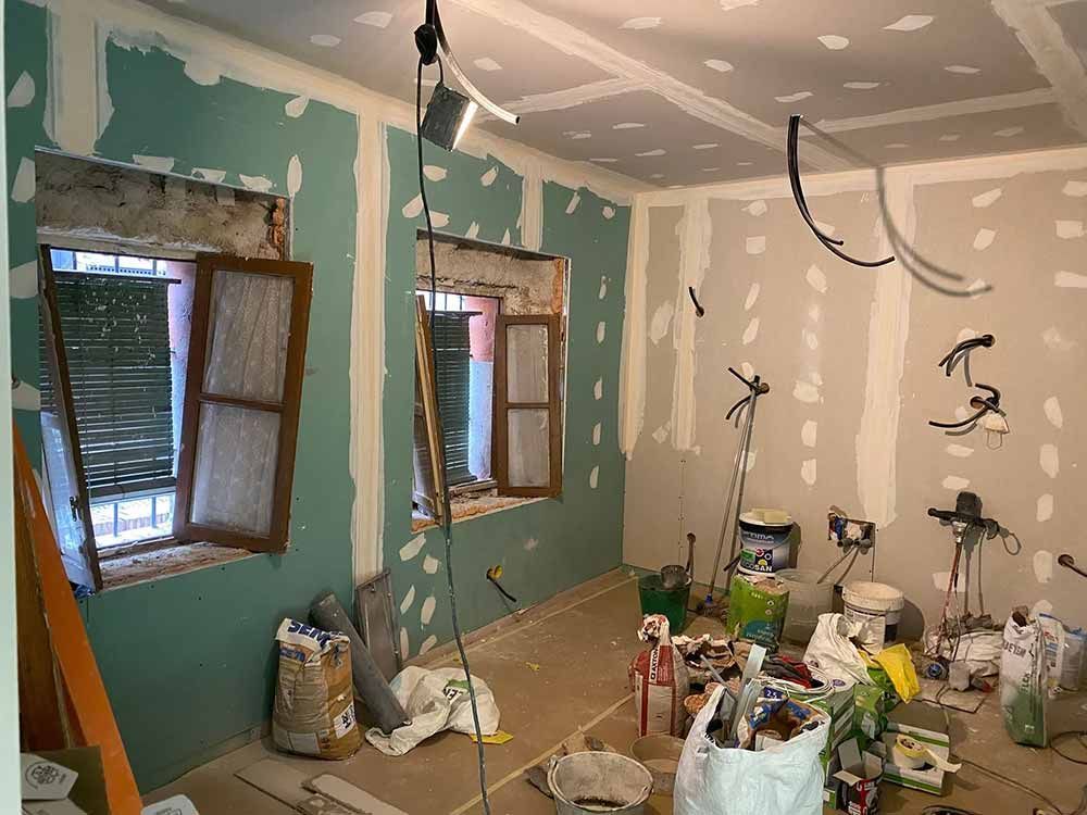 Habitación en renovación con paneles de yeso instalados. Las paredes son verdes y beige, los marcos de las ventanas son marrones. Hay herramientas y escombros esparcidos.