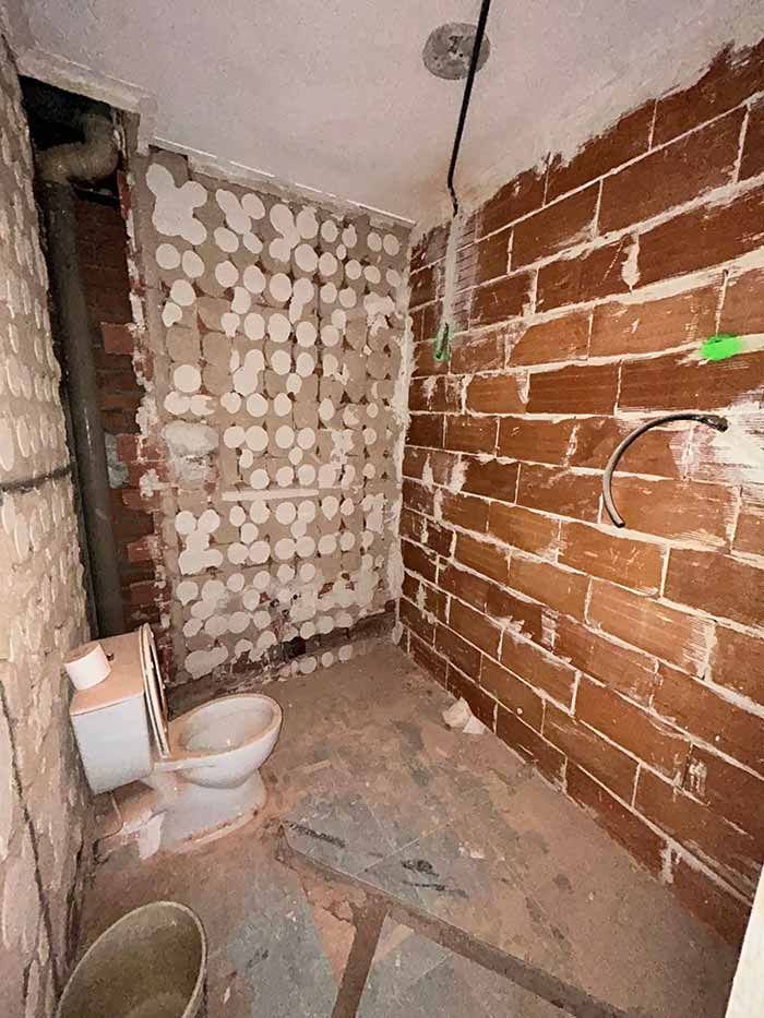 Baño en construcción: pared de ladrillo a la vista, inodoro, plomería, escombros y yeso roto.