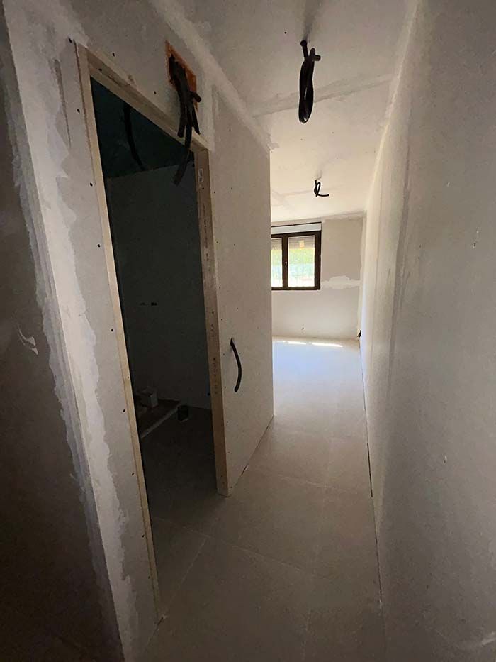 Pasillo en construcción con puerta a la izquierda y una habitación con ventana al fondo. Se ven paneles de yeso.