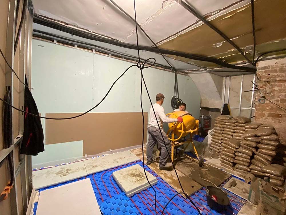 Dos personas mezclando hormigón en una habitación en construcción, con ladrillos a la vista, cables y aislamiento.