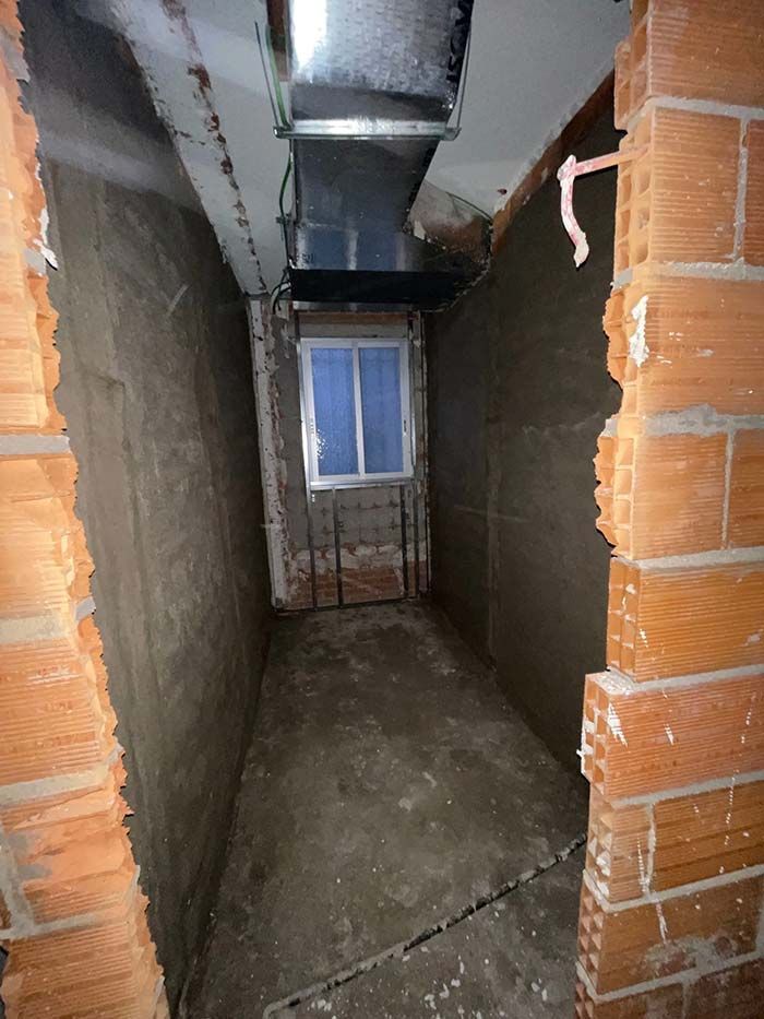 Pasillo estrecho sin terminar con ladrillo y hormigón, una ventana al final y conductos encima.