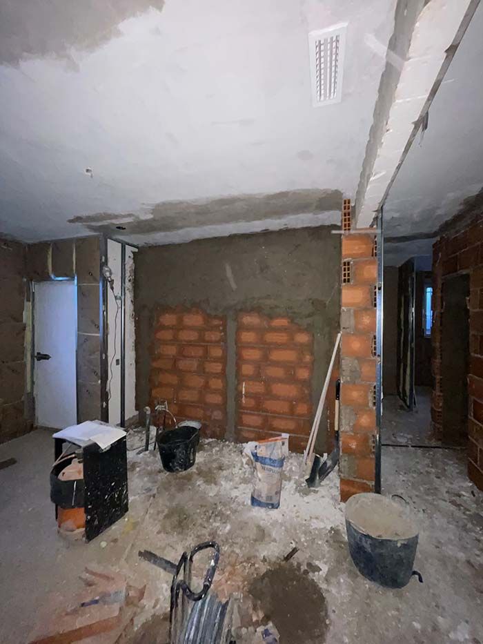 Interior en construcción: pared de ladrillo con cemento, cubetas, herramientas y ventilación de techo a la vista.
