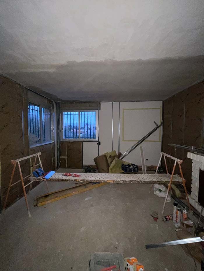 Habitación en renovación; paneles de yeso y aislamiento visibles. Caballetes que sostienen las tablas. Suelo polvoriento. Ventanas con vista al exterior.