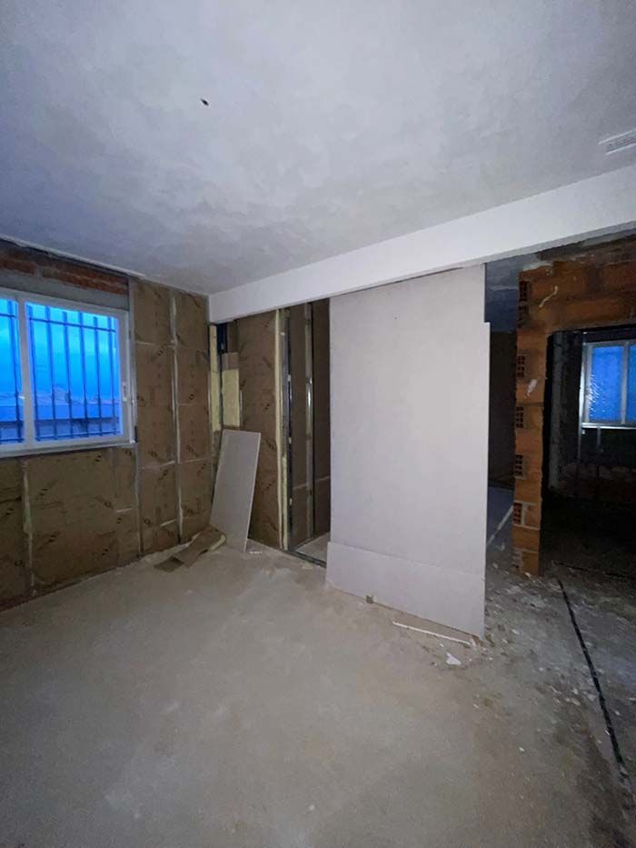 Habitación interior en construcción con aislamiento expuesto, paneles de yeso, ventanas y paredes de ladrillo.