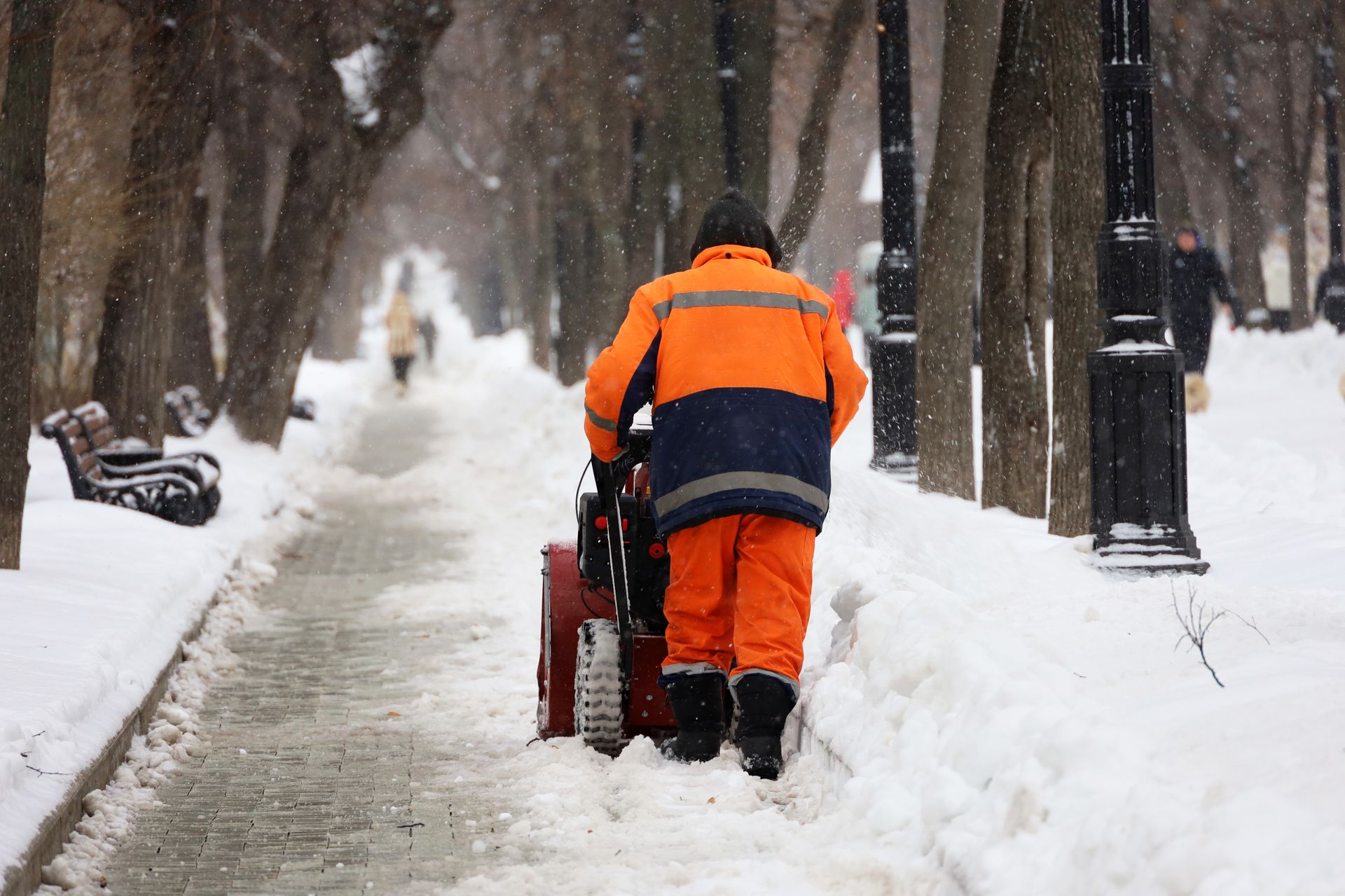 Ein Arbeiter in leuchtend orangefarbener Winterkleidung räumt mit einer roten Schneefräse Schnee von einem Gehweg.