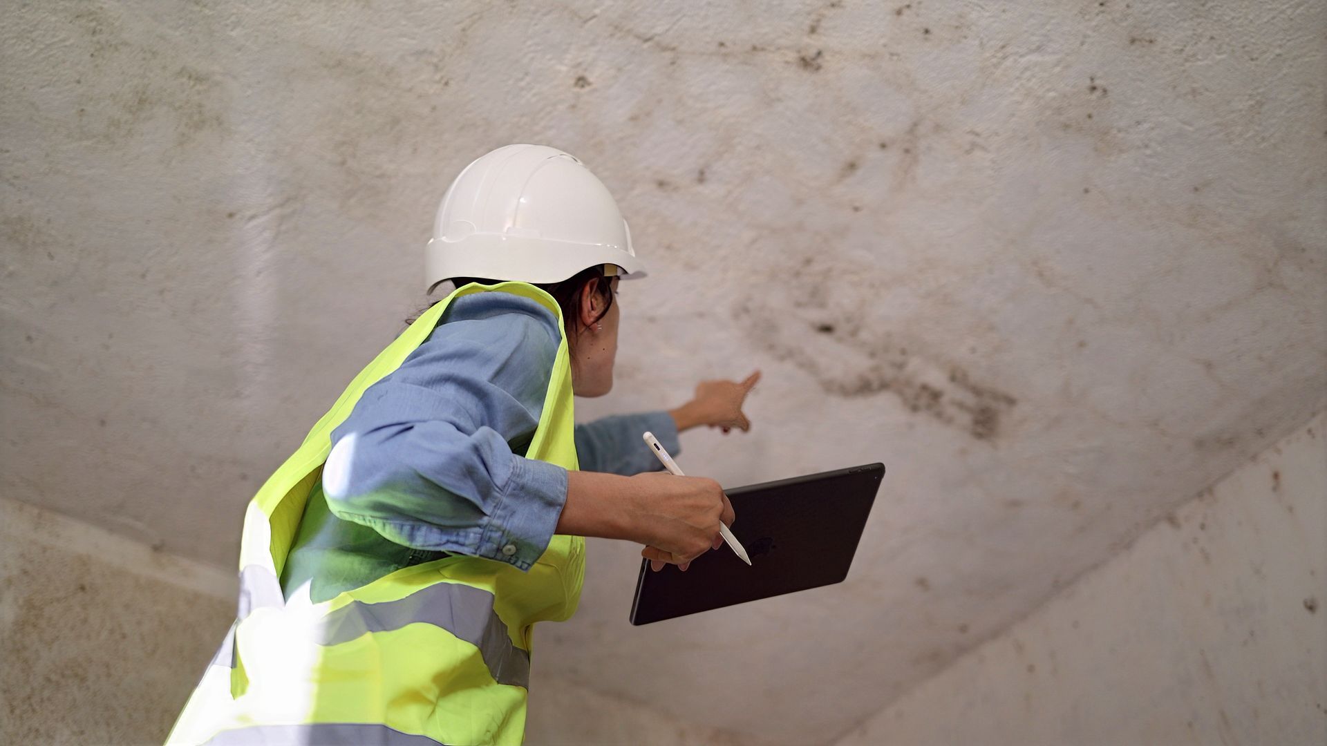 Un ouvrier portant un casque et un gilet de sécurité inspecte un plafond en béton à l'aide d'un bloc-notes.