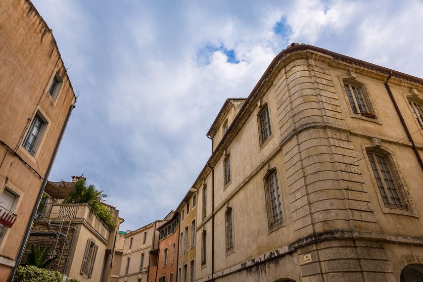 Rue historique étroite bordée de hauts bâtiments en pierre beige sous un ciel bleu nuageux.
