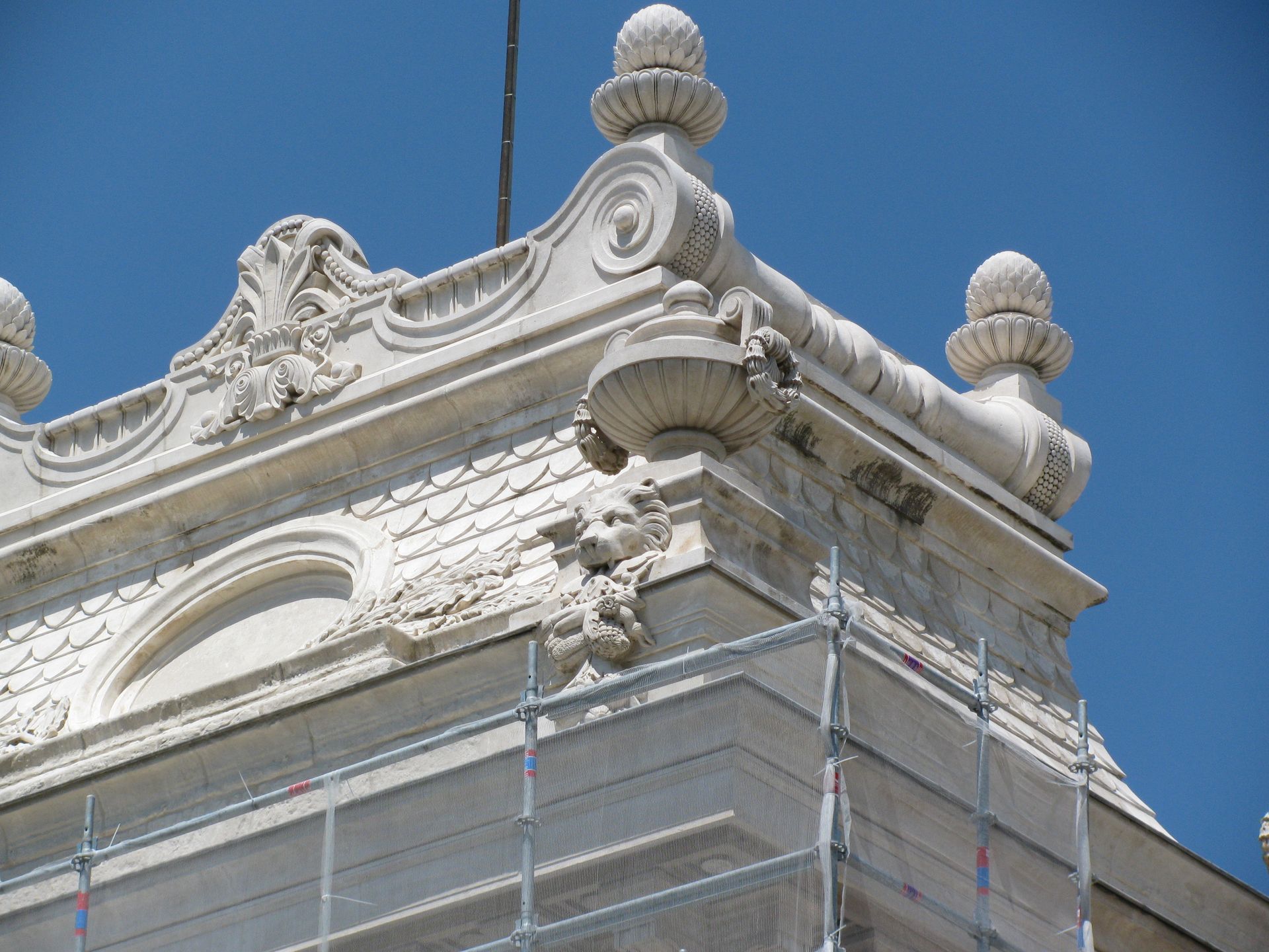 Angle d'un bâtiment blanc orné, avec des détails décoratifs sur le toit, se détachant sur un ciel bleu, avec des échafaudages en dessous.