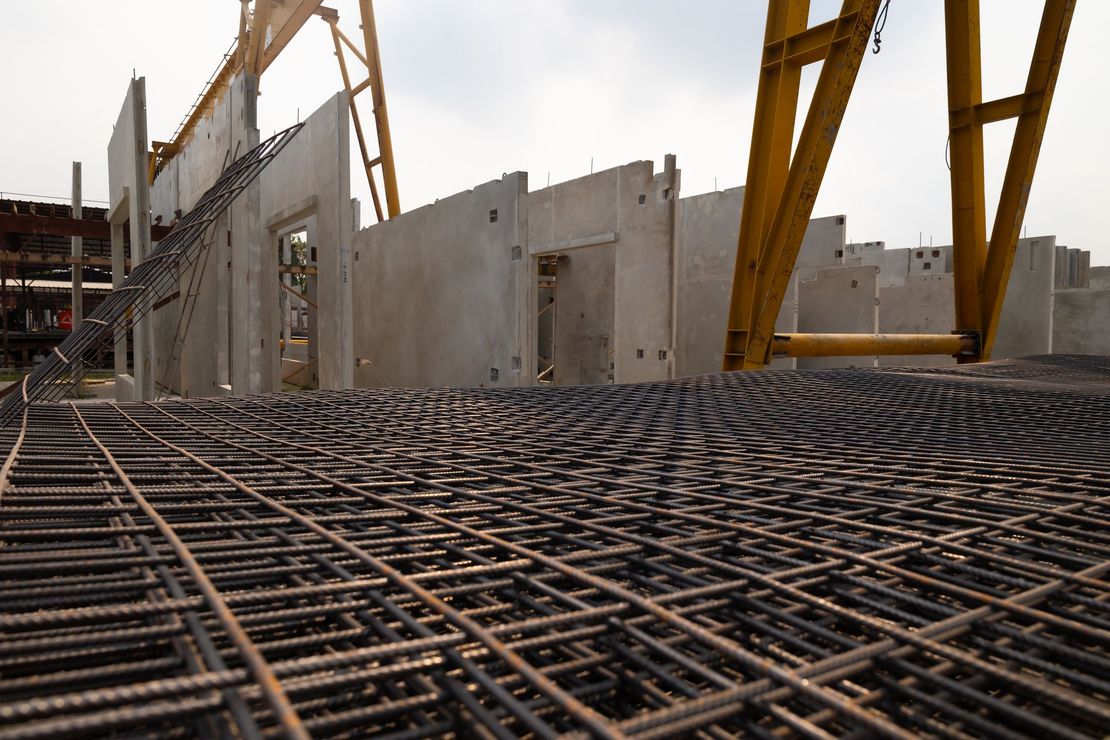 Chantier de construction avec treillis d'armatures en acier, murs en béton et grues jaunes sous un ciel nuageux.