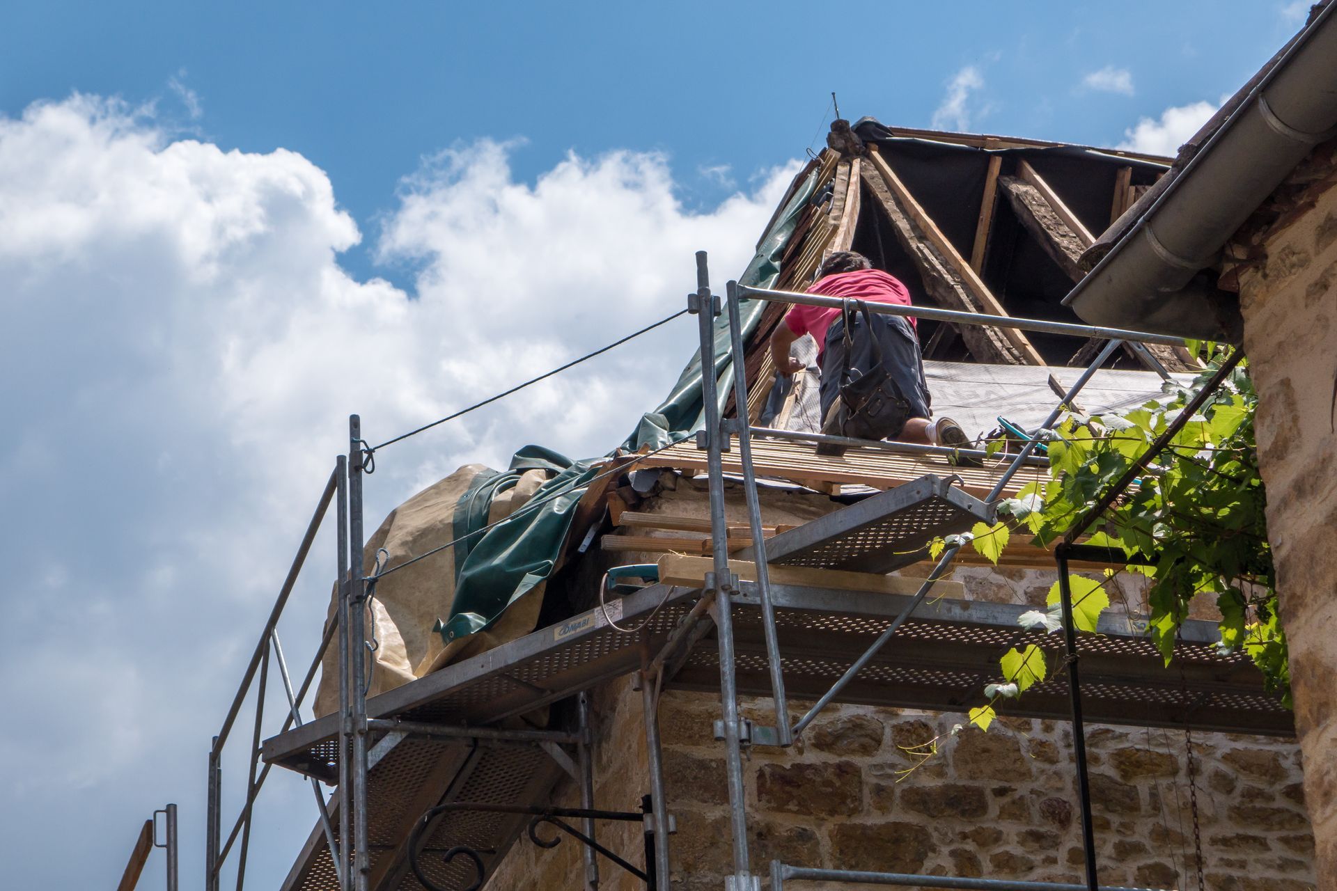 Des couvreurs réparent une maison en pierre sur des échafaudages sous un ciel bleu.