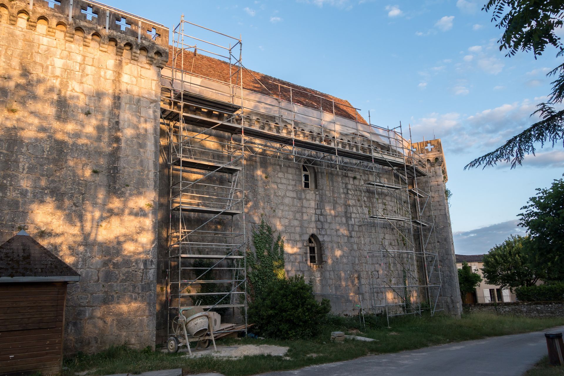 Bâtiment en pierre en rénovation, entouré d'échafaudages et baigné par la douce lumière du soleil, près d'une route et d'arbres.