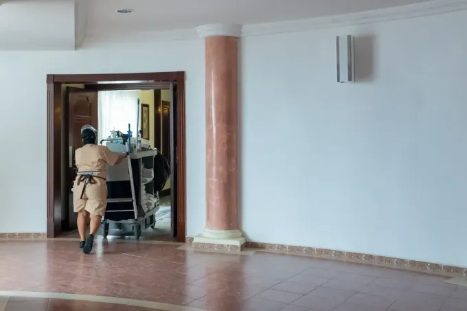 Una persona uniformada empuja un carrito a través de una puerta abierta, paredes blancas y una columna decorativa.