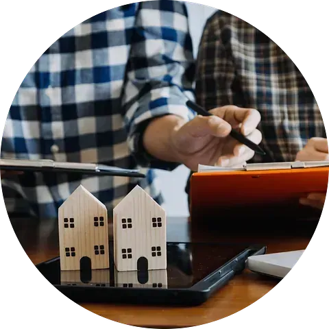 Dos casas de madera en miniatura se encuentran sobre una tableta junto a personas que revisan documentos.