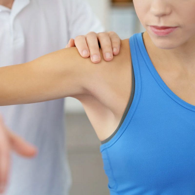 A una mujer que lleva una camiseta azul sin mangas un médico le examina el hombro.