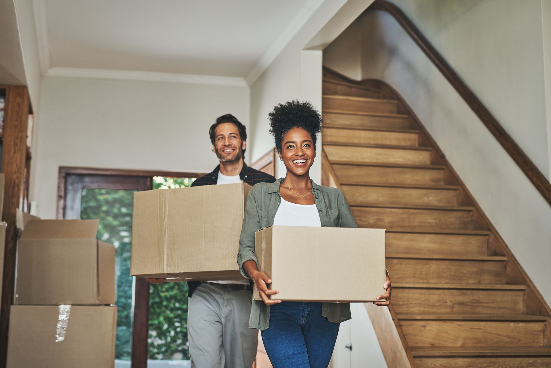 Un couple transportant des cartons de déménagement dans sa nouvelle maison, souriant près d'un escalier.