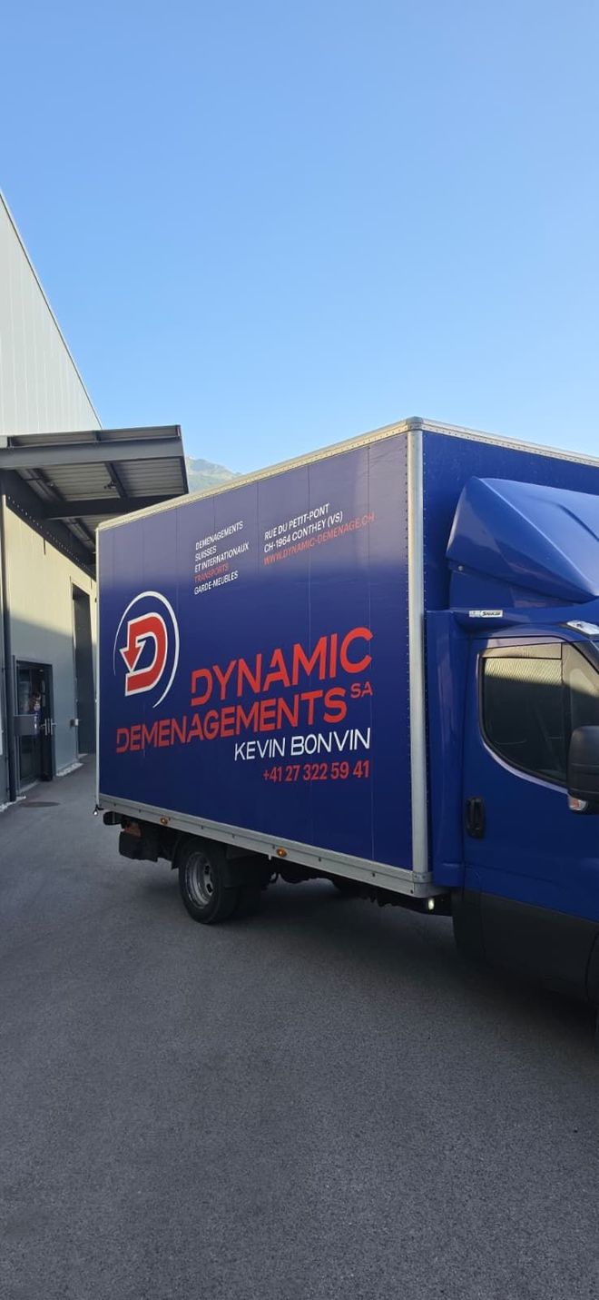 Un camion bleu est garé dans un garage-entrepôt aux hauts plafonds, avec des outils à proximité.
