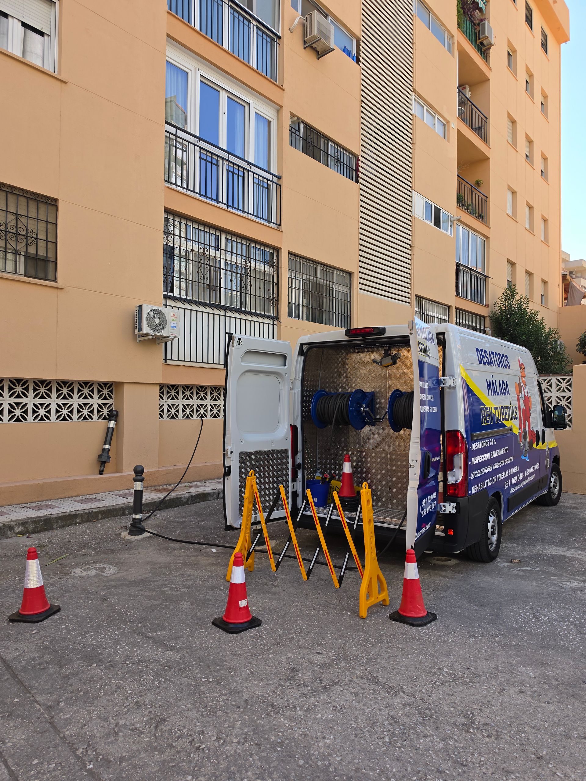 Furgoneta de mantenimiento aparcada junto a un edificio de apartamentos con conos de seguridad y equipos en el exterior.