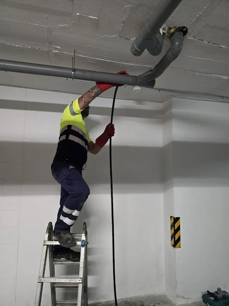 Trabajador subido a una escalera pintando o reparando una tubería del techo en un estacionamiento.