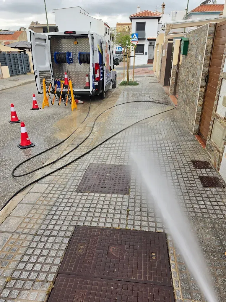 Camión de limpieza viaria lavando una acera con un chorro de agua a alta presión y conos que delimitan la zona de trabajo.